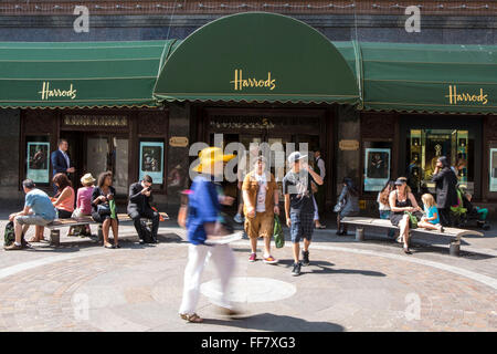 I turisti amanti dello shopping uscire dalla famosa in tutto il mondo retail store Harrods attraverso la porta 5 nella zona di Knightsbridge, Londra, Regno Unito. Alcuni altri clienti sedersi sulle panchine al di fuori del negozio godendo il sole. Foto Stock