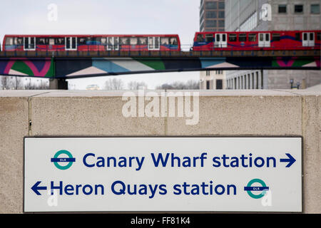 Un cartello stradale per Canary Wharf e Heron Quays DLR al di fuori delle stazioni stazione metropolitana di Canary Wharf a Londra, Inghilterra, Regno Unito. Un Docklands Light Railway (DLR) treno attraversa su un ponte sul Dock per mezzo del fiume Tamigi. La DLR è un sistema automatizzato di luce del sistema della metropolitana ed è parte del trasporto per Londra. Foto Stock