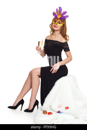 Giovane donna in nero abito da sera e un bicchiere di champagne. Il carnevale maschera sul viso. Sedersi sulla pelliccia bianca. Vacanza e il concetto di partito. Foto Stock