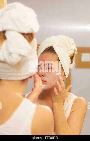 Donna di applicare la maschera idratante crema per la pelle sulla faccia guardando nello specchio del bagno. Ragazza prendersi cura della sua carnagione balsamo di stratificazione Foto Stock