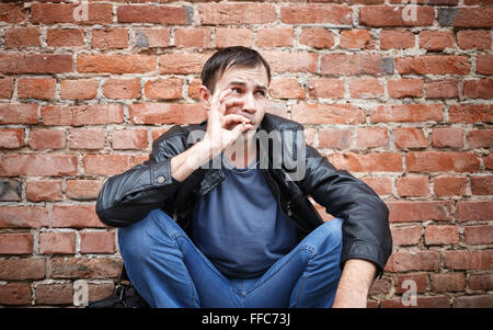 L'uomo squatting e fumare una sigaretta. Fumatori bad boy contro il vecchio muro di mattoni in background. Foto Stock