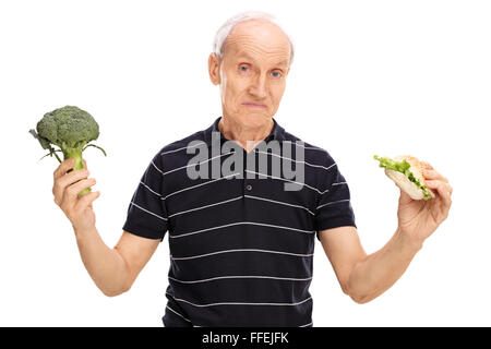 Indeciso senior uomo in possesso di un pezzo di broccoli in una mano e un panino in altri isolati su sfondo bianco Foto Stock