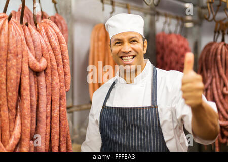 Allegro media età del macellaio dando il pollice in su contro wors appesi in macelleria Foto Stock