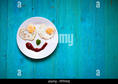 La prima colazione, uova fritte in forma di cuori, Smile, buon umore, mattino luminoso blu sullo sfondo di legno Foto Stock
