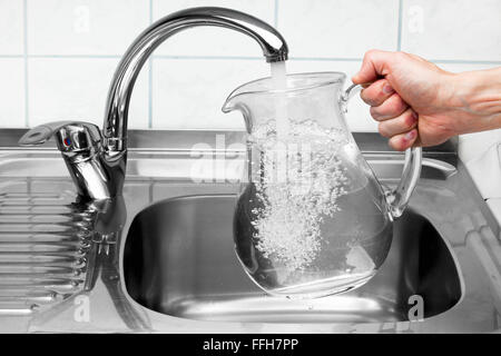 Mano che tiene una brocca di acqua versata dal rubinetto di cucina. Foto Stock