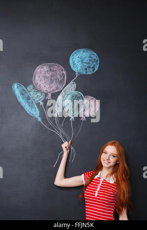 Felice attraente redhead giovane donna in piedi e tenendo palloncini disegnati su sfondo blackboard Foto Stock