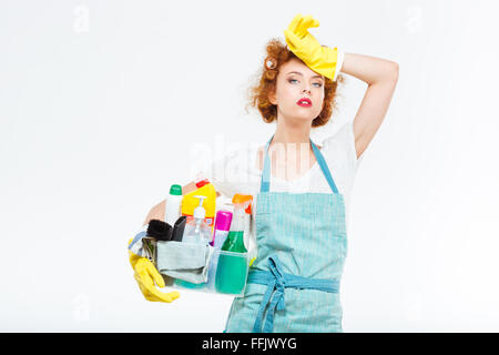 Stanco attraente giovane donna in giallo i guanti e il grembiule blu permanente e la scatola di contenimento con detergenti su sfondo bianco Foto Stock