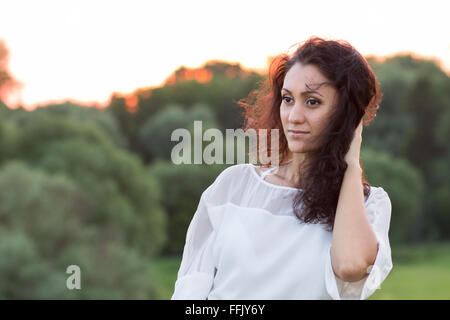 Giovane donna felice con il nero capelli crespi in piedi all'aperto contro boschi in serata. Ritratto di ispanici ragazza sorridente in wh Foto Stock