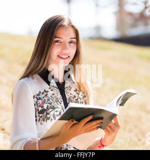 I giovani europei studentessa con la cartella di lavoro in all'aperto sul giorno caldo e soleggiato. Sorridente ragazza con libro seduti sulla collina di erba Foto Stock