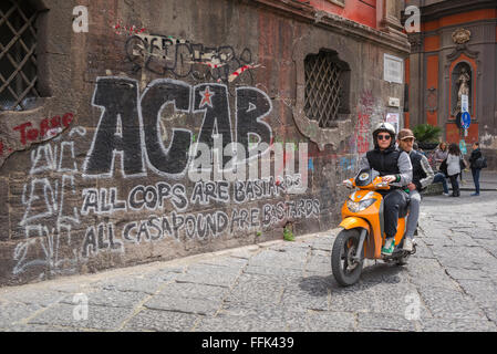 Via Napoli, vista di un paio di ragazzi napoletani che cavalcano uno scooter passando davanti a un muro graffiato in Piazza San Domenico, nel centro di Napoli, Italia. Foto Stock