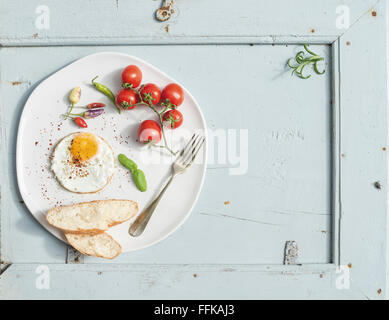 Set colazione. Uovo fritto, fette di pane, pomodori ciliegini, peperoncino e erbe sul piatto di portata in ceramica bianca sulla luce blu bac in legno Foto Stock