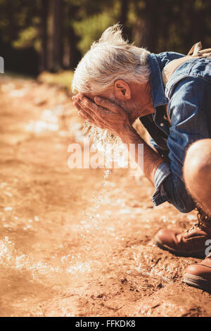 Lato ritratto di un uomo anziano lavando il suo volto nel lago. Maschio maturo escursionista getting fresco con acqua di lago. Foto Stock