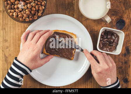 Donna diffusione di nocciola crema al torrone su fetta di pane, vista dall'alto di mani femminili al tavolo della cucina. Foto Stock