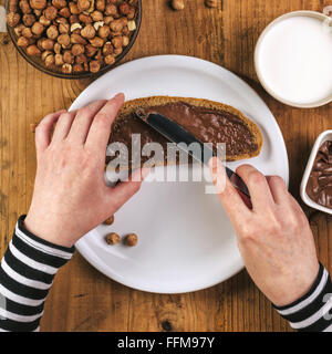 Donna diffusione di nocciola crema al torrone su fetta di pane, vista dall'alto di mani femminili al tavolo della cucina. Foto Stock