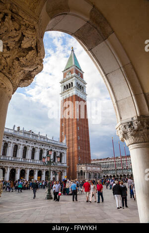 Vista di St Marks torre sulla piazza San Marco, Venezia, attraverso un arco del Palazzo dei Dogi con molti turisti che si godono il sole Foto Stock