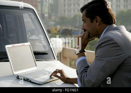 Commerciante indiano in cappotto grigio utilizzando laptop sul cofano della vettura contro edifici aziendali di Pune City , Maharashtra Foto Stock