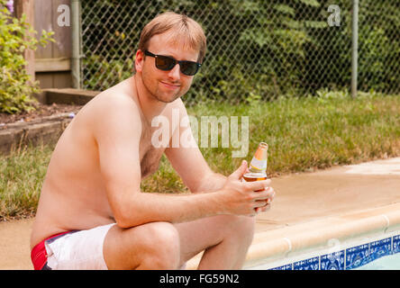 Giovani caucasici maschio adulto seduto sul bordo di una piscina in un giorno caldo e soleggiato gustando una bevanda fresca. Foto Stock
