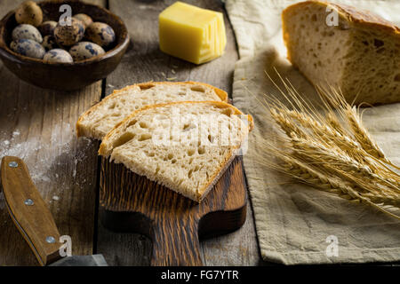 Pane di pasta acida e un pezzo di formaggio, uova di quaglia, dorate spighe di grano e il coltello sulla tavola in legno rustico. Piatti rustici still life Foto Stock