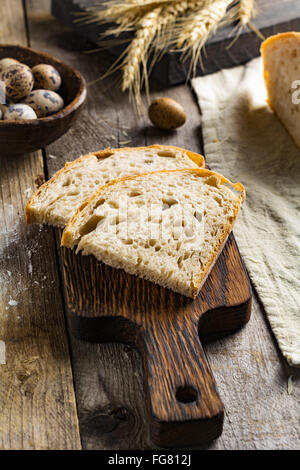 Pane di pasta acida e un pezzo di formaggio, uova di quaglia, dorate spighe di grano e il coltello sulla tavola in legno rustico. Piatti rustici still life Foto Stock