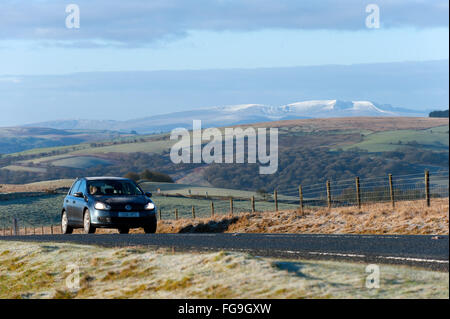 Builth Wells, Powys, Regno Unito. 18 Febbraio, 2016. Automobilista aziona attraverso un gelido paesaggio sulla B4520 road (il Brecon Road) in alto la brughiera di Mynydd Epynt intervallo vicino Builth Wells. Vette innevate del Parco Nazionale di Brecon Beacons sono visibili in lontananza. Credito: Graham M. Lawrence/Alamy Live News. Foto Stock