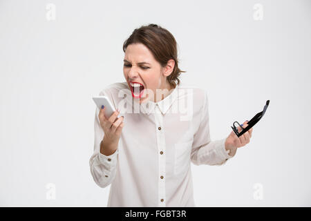 Arrabbiato irritato young business woman tenendo il telefono cellulare e bicchieri e gridando su sfondo bianco Foto Stock
