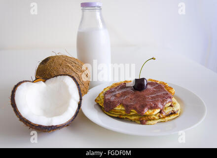 La colazione e il brunch frittelle di cocco pasto con crepe aperte coco, bottiglia di vetro di latte e ciliegia su sfondo bianco. Pila di Foto Stock