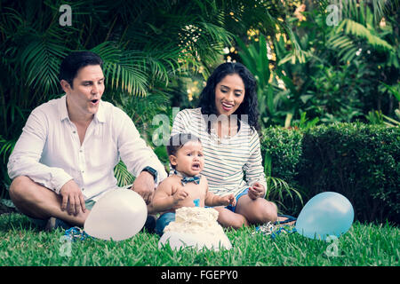 Padre, madre e neonato bambino seduto nell'erba per celebrare il 1. compleanno con torta e palloncini in giardino. Foto Stock