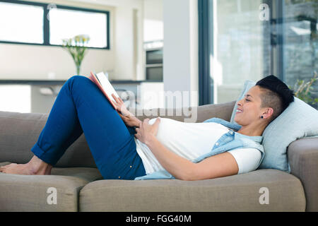 Donna incinta a leggere un libro mentre sdraiato sul divano Foto Stock