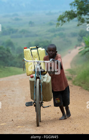 Ragazzo spingendo una bicicletta laden con pieno di acqua contenitori lungo una strada polverosa, Uganda. Foto Stock