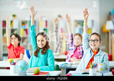 Felice gli alunni seduti a scrivanie e alzando le mani alla lezione Foto Stock