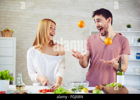 Giocoso giovane juggling agrumi con sua moglie stupito per la cottura nelle vicinanze Foto Stock