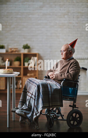 Senior uomo sulla sedia a rotelle seduto di fronte a tavola con torta di compleanno Foto Stock