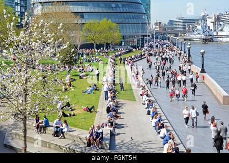 Sole primaverile per i turisti e gli impiegati di godere lungo la riva sud accanto al Fiume Tamigi a City Hall Southwark più in Londra England Regno Unito Foto Stock