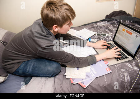 White ragazzo adolescente seduto sul suo letto con un computer portatile e un sacco di libri scolastici come egli rivede per esami GCSE e rende nota per la sua scuola e compiti. Foto Stock