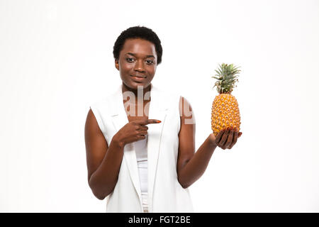 Felice attraente americano africano giovane donna holding ananas e puntando su di esso su sfondo bianco Foto Stock