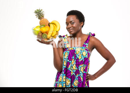 Allegro attraente americano africano giovane donna in bright sundress permanente e la holding ciotola di vetro con frutti su bianco backg Foto Stock