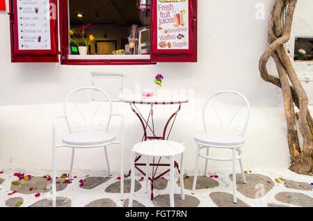 Un coffee shop a Chora sull'isola greca di Mykonos, Grecia in un tradizionale lastricate di ciottoli con vicolo rosso vetri di finestre, sedie, t Foto Stock