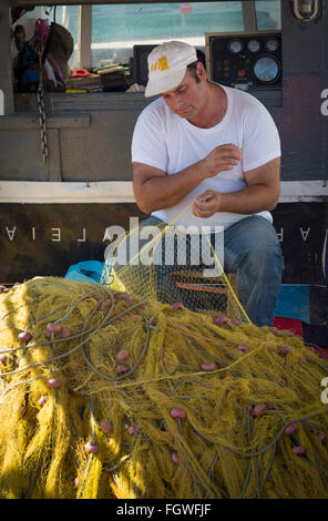 Volos, Tessaglia, Grecia. Un pescatore seduto sulla sua barca ormeggiata in porto mende le sue reti. Foto Stock