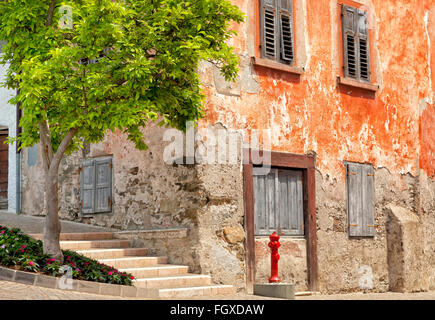 Vecchio rosso storica casa d'angolo con età compresa tra pareti e finestre di otturatore, sulla strada di ciottoli, con acqua rossa idrante e albero nella parte anteriore Foto Stock