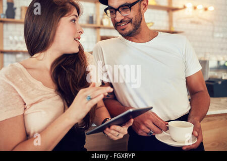 Ritratto di felice coppia giovane presso il cafe contatore con la discussione su una tazza di caffè. Donna che mantiene una tavoletta digitale e uomo wi Foto Stock