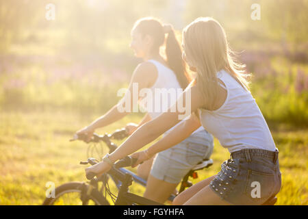 Due giovani graziosi felice belle ragazze che indossano bianco casual canotte e gli shorts in denim biciclette equitazione nel parco in bright Foto Stock