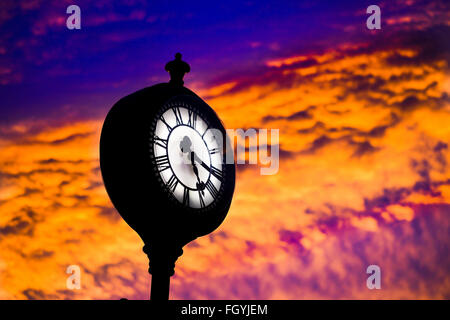 Orologio pubblico contro un forte tramonto che mostra il tempo su un numero romano faccia del trimestre passato cinque. Foto Stock