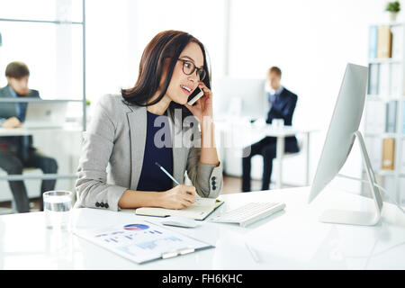 Occupato segretario parlando al cellulare e rendere note in notebook mentre guardando il monitor Foto Stock