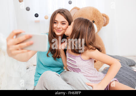 Due graziose sorelle sorridente baciare e rendendo selfie in camera per bambini Foto Stock