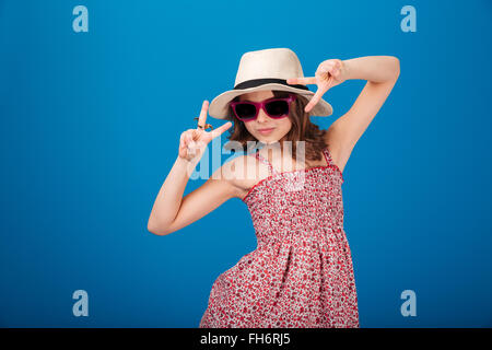 Carino giocoso bambina in hat, sundress e occhiali da sole che mostra segno di vittoria con entrambe le mani su sfondo blu Foto Stock