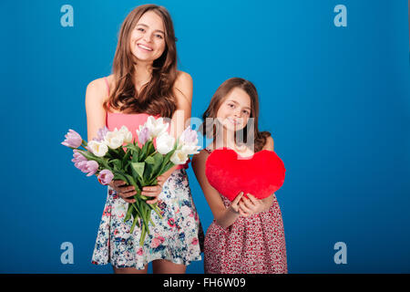 Due allegro carino sorelle holding bouquet di fiori e di cuore su sfondo blu Foto Stock