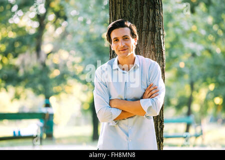 Uomo adulto in maglietta blu appoggiato su di un albero in posizione di parcheggio Foto Stock