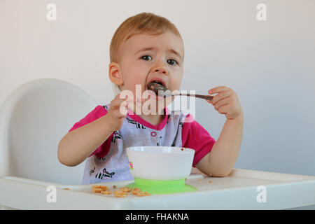 Poco sorridente baby si siede sul seggiolone e mangia porridge sulla piastra. Foto Stock