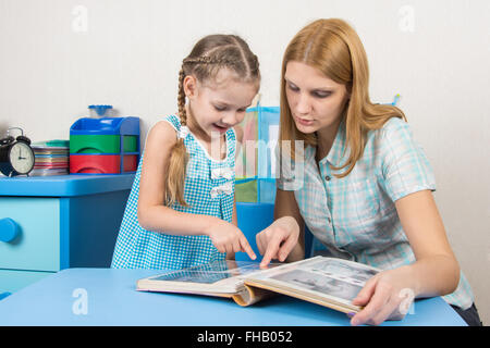 Adulto giovane bella ragazza mostra cinque anni di ragazza in album di foto seduti al tavolo per bambini a casa Foto Stock