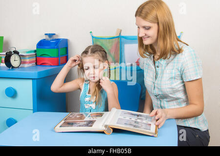 Adulto giovane bella ragazza mostra cinque anni di ragazza in album di foto seduti al tavolo per bambini a casa Foto Stock